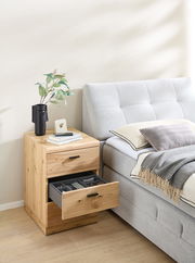 Holz-Rollcontainer mit zwei Schubladen, eine geöffnet, neben einem grauen Bett. Perspektive von vorne.