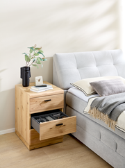 Holz-Rollcontainer mit zwei Schubladen, eine geöffnet, neben einem grauen Bett. Perspektive von vorne.