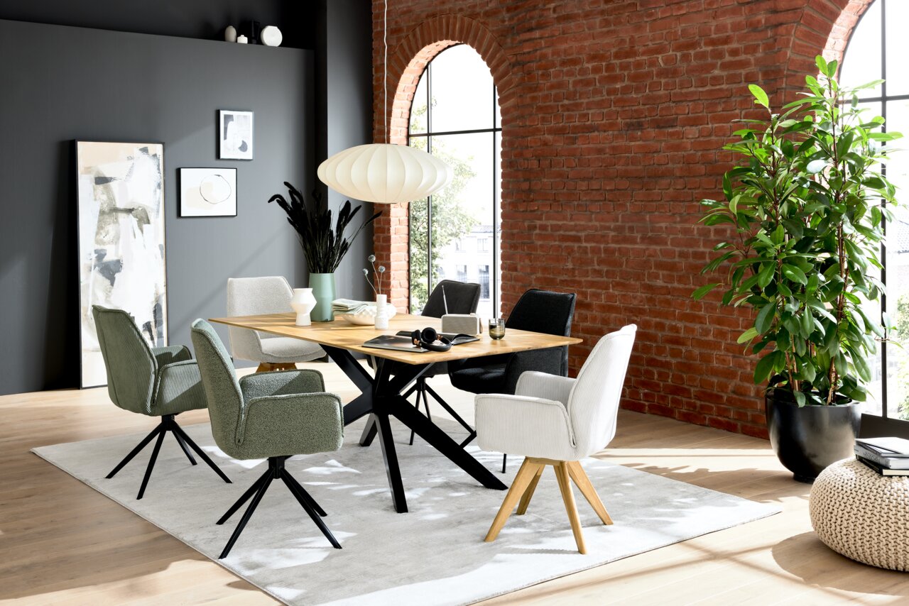 Moderner Esstisch mit Holzplatte und schwarzen Metallbeinen, umgeben von verschiedenen Stühlen in einem stilvollen Raum mit Backsteinwand und großen Fenstern, seitliche Perspektive.