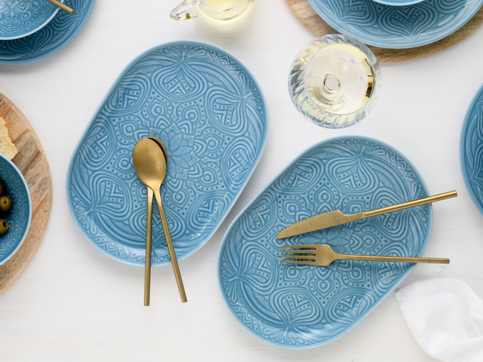 Draufsicht auf zwei ovale, blaue Teller mit geprägtem Mandala-Muster auf weißem Tisch, mit goldfarbenem Besteck und einem Weinglas dekoriert