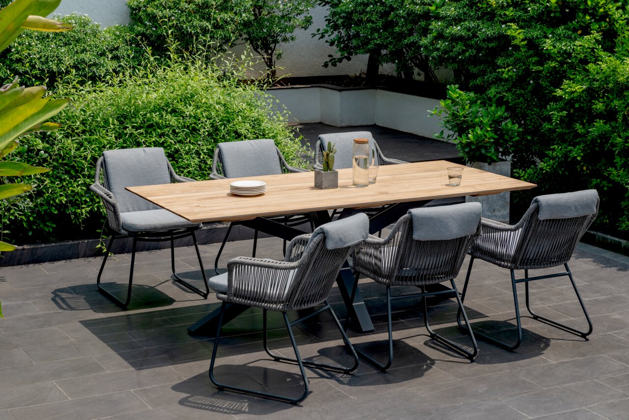 Amatio Fauteuil CIRCLE Moderne houten eettafel voor buiten met een zwart metalen onderstel, omringd door zes grijze tuinstoelen met gevlochten koord op een terras; perspectief: licht schuin van voren op ooghoogte.