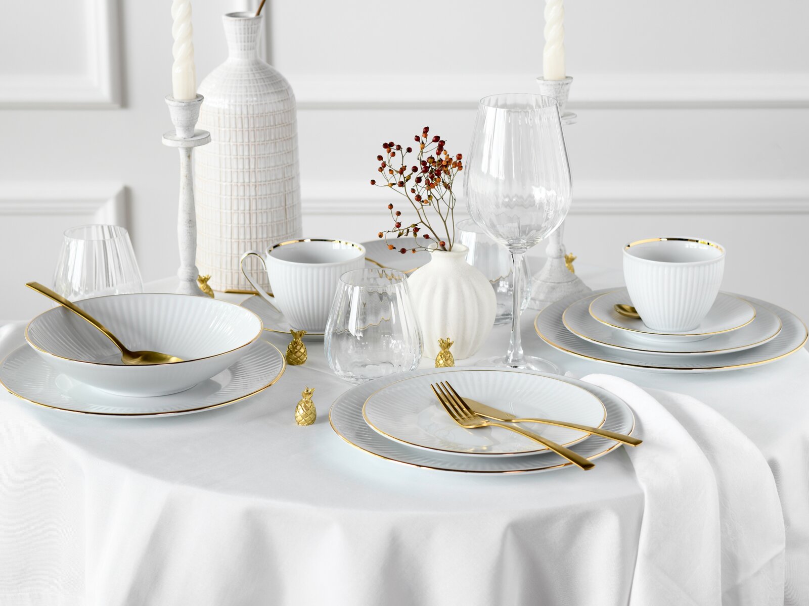 Elegantes weißes Tafelservice mit feiner Rillenstruktur und Goldrand, gedeckter Tisch mit goldfarbenem Besteck, Weinglas und Dekovasen; Perspektive: leicht erhöhte Frontansicht.
