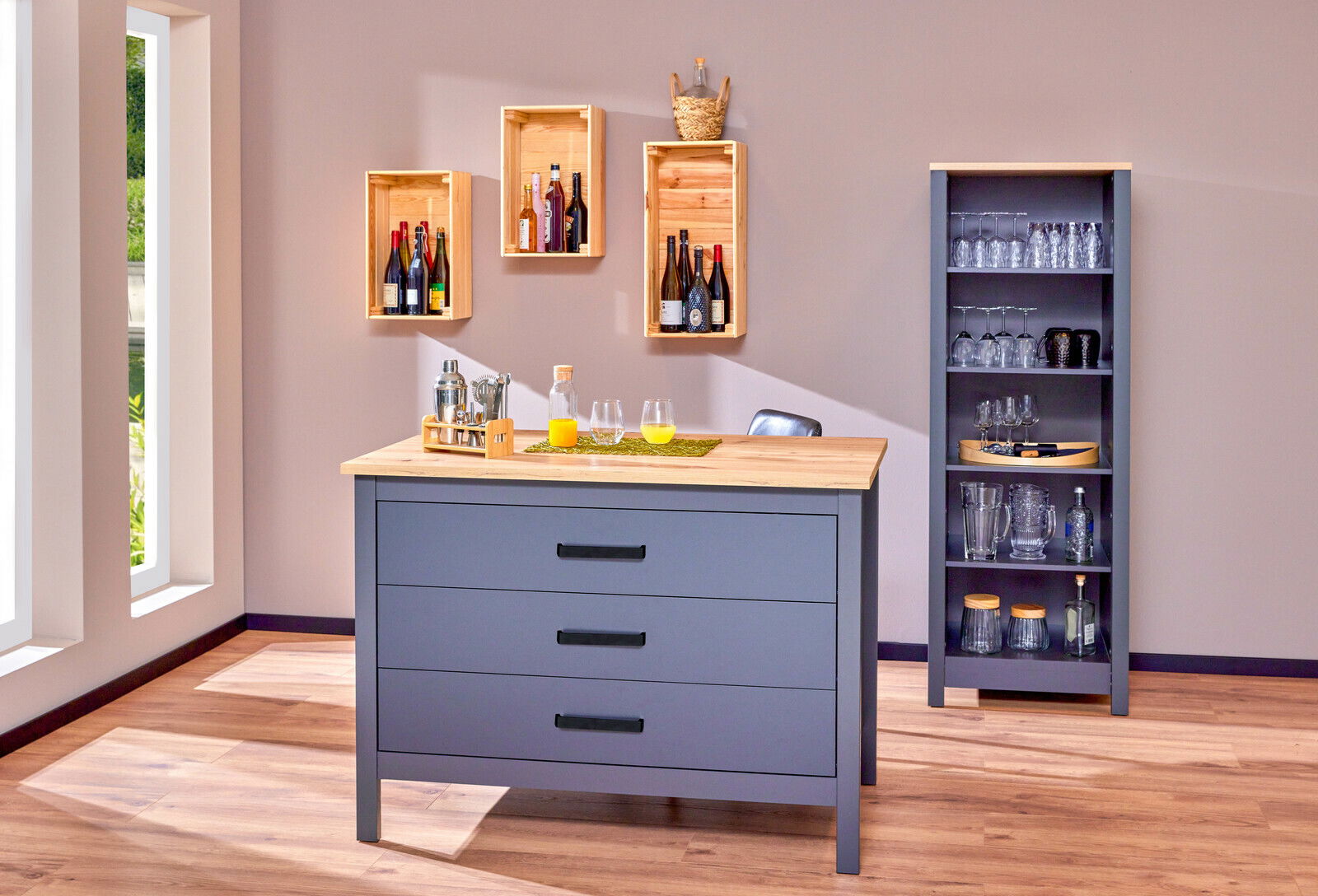 Kücheninsel mit drei Schubladen in grauer Farbe und Holzarbeitsplatte, frontal fotografiert. Im Hintergrund sind Regale mit Gläsern und Flaschen zu sehen.