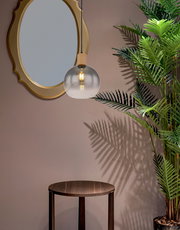 Hängeleuchte mit rundem, transparentem Glasschirm und Holzdetail, von der Seite betrachtet, neben einem runden Spiegel und einer Pflanze.