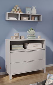Kommode und Wandregal in einem Babyzimmer, frontal fotografiert. Auf der Kommode stehen Spielzeug und Bücher, während das Regal dekorative Gegenstände enthält.