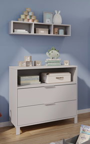 Kommode und Wandregal in einem Babyzimmer, frontal fotografiert. Auf der Kommode stehen Spielzeug und Bücher, während das Regal dekorative Gegenstände enthält.