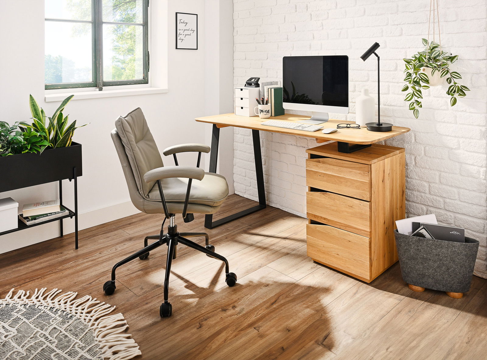 Moderner Schreibtisch aus Holz mit schwarzem Metallgestell, seitlich von einem Bürostuhl und Pflanzen umgeben, in einem hellen Raum mit Fenster und weißer Ziegelwand.