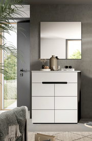 Modernes weißes Sideboard/Kommode mit schwarzer Griffleiste vor Betonwand, darüber rechteckiger Wandspiegel; dekoriert mit Vasen und Büchern, Perspektive: frontale Ansicht auf Augenhöhe.