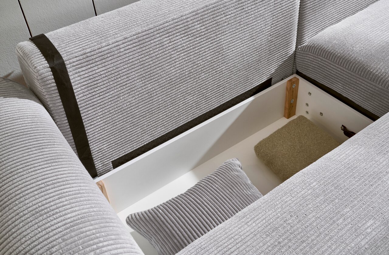 Nahaufnahme einer grauen Eckcouch mit Stauraum, geöffnet, mit Kissen und Decke. Perspektive von oben.