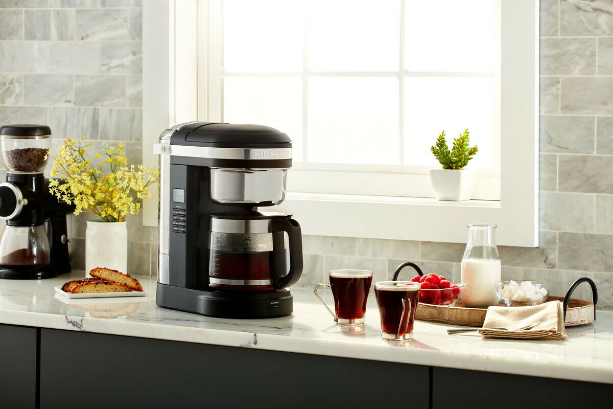 Kaffeemaschine in onyx schwarz auf einer Küchenarbeitsplatte, umgeben von zwei Tassen Kaffee, einer Karaffe Milch, einer Schale mit Himbeeren und Zuckerwürfeln, mit Blick aus einem Fenster im Hintergrund.