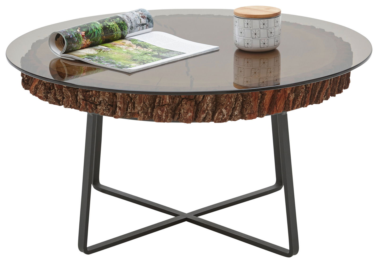 Runder Couchtisch mit Glasplatte und rustikalem Holzrand, auf schwarzem Metallgestell, aus seitlicher Perspektive fotografiert. Auf dem Tisch liegen eine aufgeschlagene Zeitschrift und ein kleiner Behälter mit Holzdeckel.
