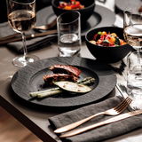 Eleganter Speiseteller aus schwarzem Material mit feiner Struktur, präsentiert in einer stilvollen Tischszene mit Gourmet-Essen und edlem Besteck, aus der Vogelperspektive.