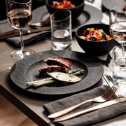 Eleganter Speiseteller aus schwarzem Material mit feiner Struktur, präsentiert in einer stilvollen Tischszene mit Gourmet-Essen und edlem Besteck, aus der Vogelperspektive.