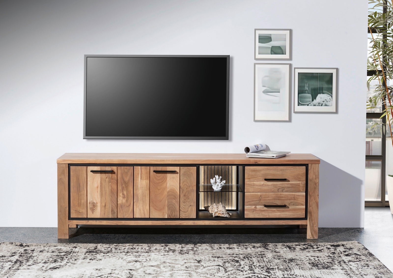 Frontale Ansicht eines Lowboards aus Holz mit zwei Türen, zwei Schubladen und einem offenen Fach mit Beleuchtung, dekoriert mit Korallen und Muscheln, vor einer weißen Wand mit einem großen Fernseher und gerahmten Bildern.