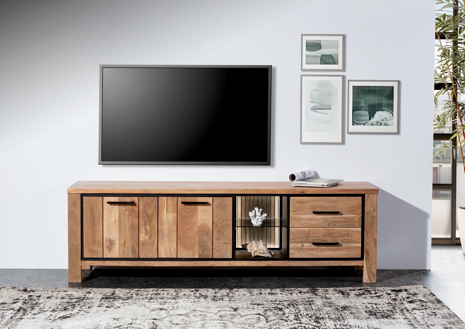 Frontale Ansicht eines Lowboards aus Holz mit zwei Türen, zwei Schubladen und einem offenen Fach mit Beleuchtung, dekoriert mit Korallen und Muscheln, vor einer weißen Wand mit einem großen Fernseher und gerahmten Bildern.