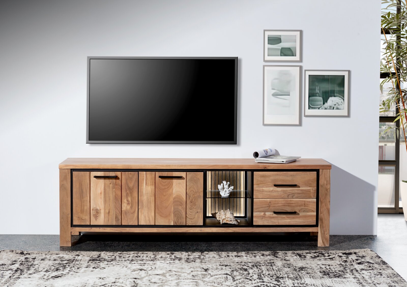 Frontale Ansicht eines Lowboards aus Holz mit zwei Türen, zwei Schubladen und einem offenen Fach mit Beleuchtung, dekoriert mit Korallen und Muscheln, vor einer weißen Wand mit einem großen Fernseher und gerahmten Bildern.