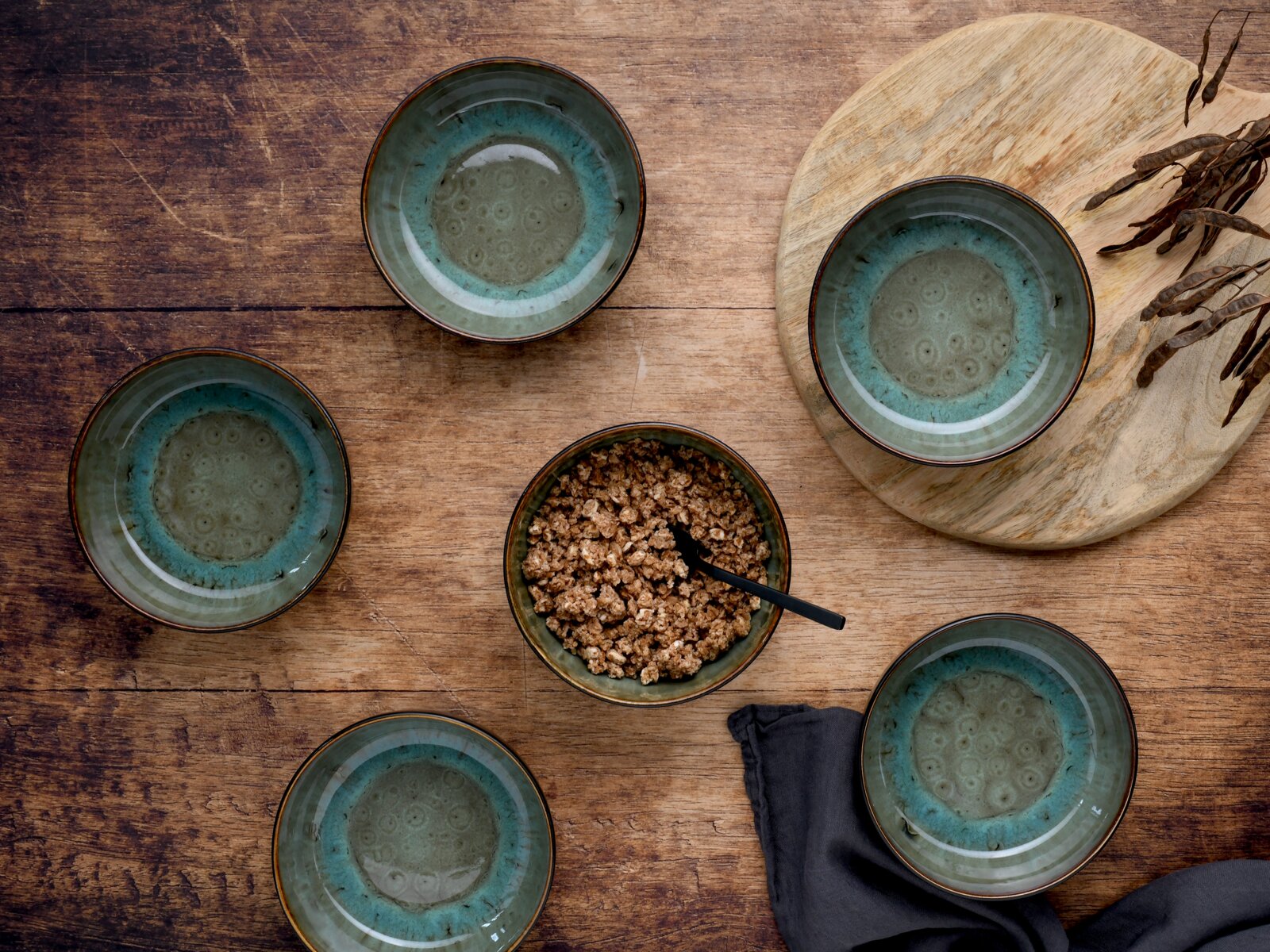 Draufsicht auf fünf grün-blau glasierte Keramikschalen auf rustikalem Holztisch, eine Schale mit Müsli und schwarzem Löffel, daneben Holzbrett und dunkles Tuch
