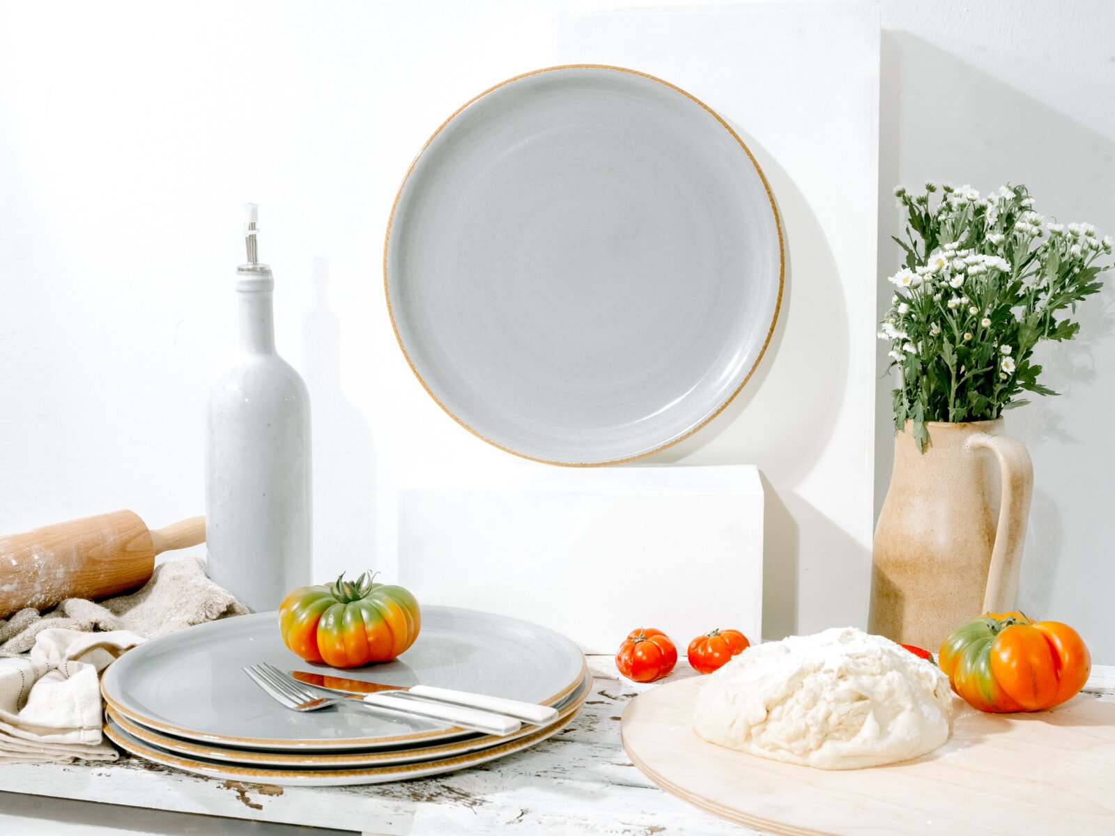 Heller grauer Keramikteller mit beigem Rand, frontal von oben (Top-Down) fotografiert, in stilisiertem Küchen-Stillleben mit weiteren Tellern, Tomaten, Teig, Ölflasche, Nudelholz und Blumen auf weißem Hintergrund.