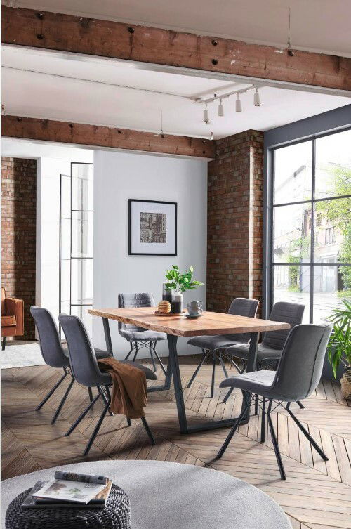 Esszimmer mit modernem Holztisch und V-Gestell, umgeben von sechs grauen Stühlen. Der Raum hat große Fenster und eine Ziegelwand, die eine helle und einladende Atmosphäre schaffen. Perspektive: seitliche Ansicht des Tisches.
