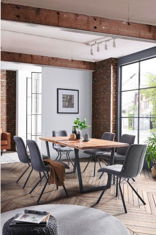 Eetkamer met een moderne houten tafel met V-vormig onderstel, omringd door zes grijze stoelen. De ruimte heeft grote ramen en een bakstenen muur, die zorgen voor een lichte en uitnodigende sfeer. Perspectief: zijaanzicht van de tafel.