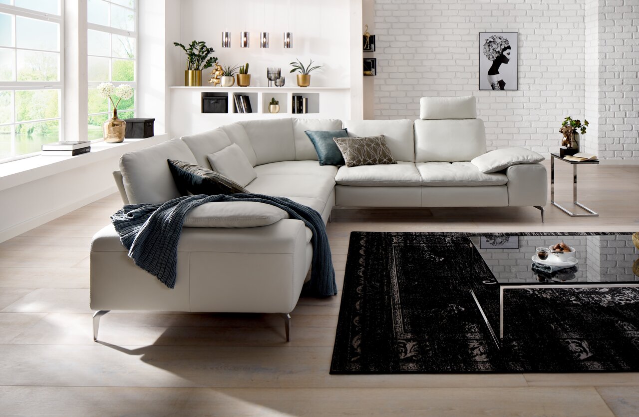 Moderne Polsterrundecke in einem hellen Wohnzimmer mit großen Fenstern, dekorativen Kissen und einem eleganten Couchtisch. Perspektive von vorne links.
