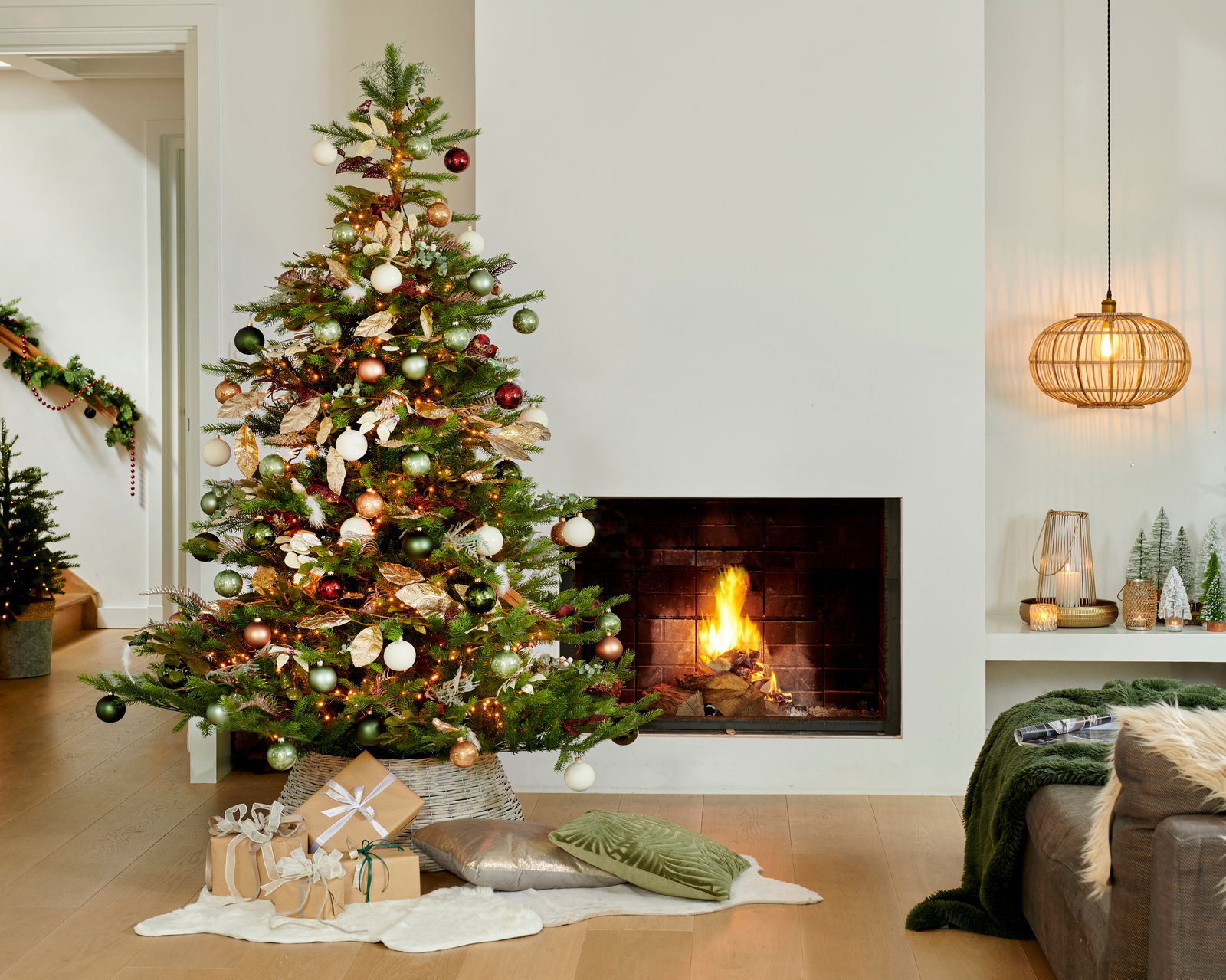Weihnachtsbaum mit bunten Kugeln und Lichtern, neben einem brennenden Kamin, aus der Perspektive des Wohnzimmers.