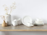 Minimalistisches weißes Porzellan-Geschirrset mit Tassen, Untertassen und Tellern auf Holztisch neben zwei Vasen mit Trockenblumen, frontal in leichter Seitenansicht
