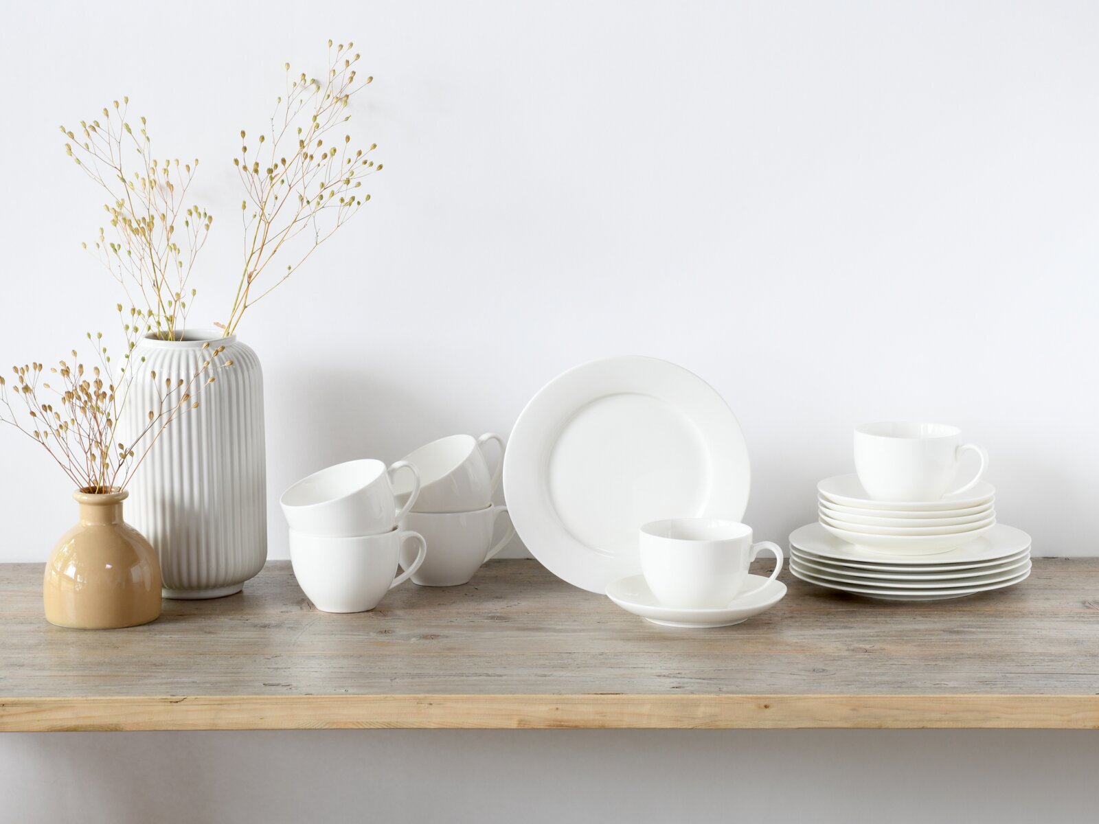 Creatable Kaffeeservice 18-tlg FLEUR ROYAL Minimalistisches weißes Porzellan-Geschirrset mit Tassen, Untertassen und Tellern auf Holztisch neben zwei Vasen mit Trockenblumen, frontal in leichter Seitenansicht