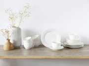 Minimalistisches weißes Porzellan-Geschirrset mit Tassen, Untertassen und Tellern auf Holztisch neben zwei Vasen mit Trockenblumen, frontal in leichter Seitenansicht