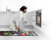 Küchenläufer mit buntem Gemüse-Design und der Aufschrift 'Natural Food', in einer modernen weißen Küche vor einem Backofen platziert. Perspektive von der Seite.