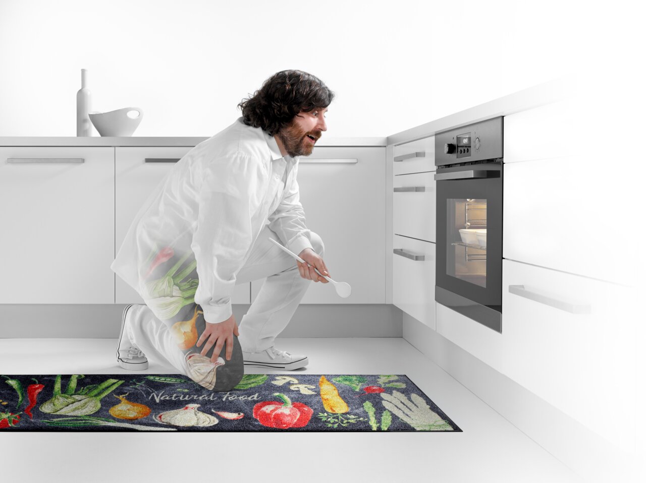 Küchenläufer mit buntem Gemüse-Design und der Aufschrift 'Natural Food', in einer modernen weißen Küche vor einem Backofen platziert. Perspektive von der Seite.