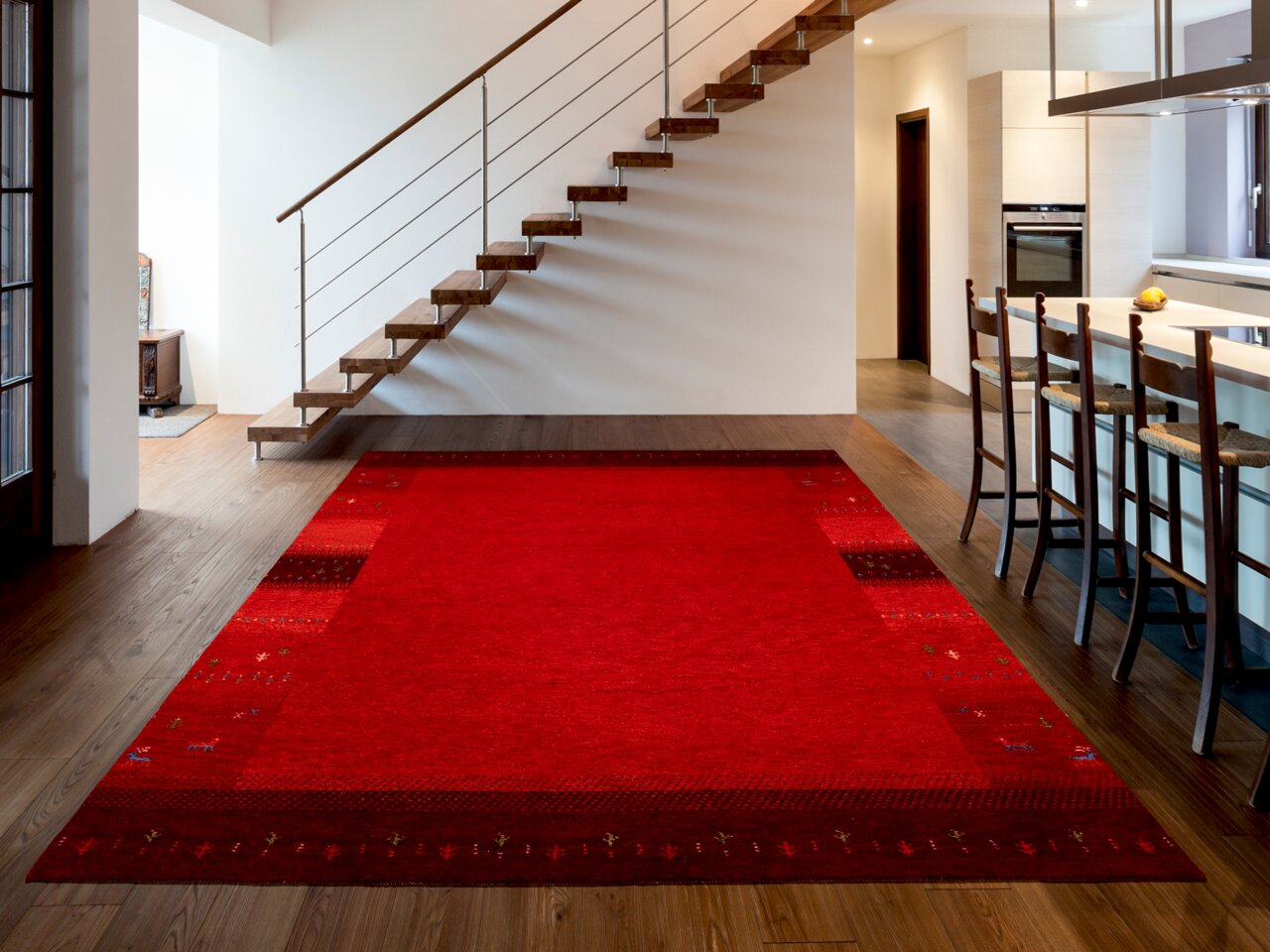 OCI Wollteppich INTENSE Roter Wollteppich in einem modernen Wohnzimmer mit Holztreppe und offener Küche, aus der Perspektive des Raumes fotografiert.