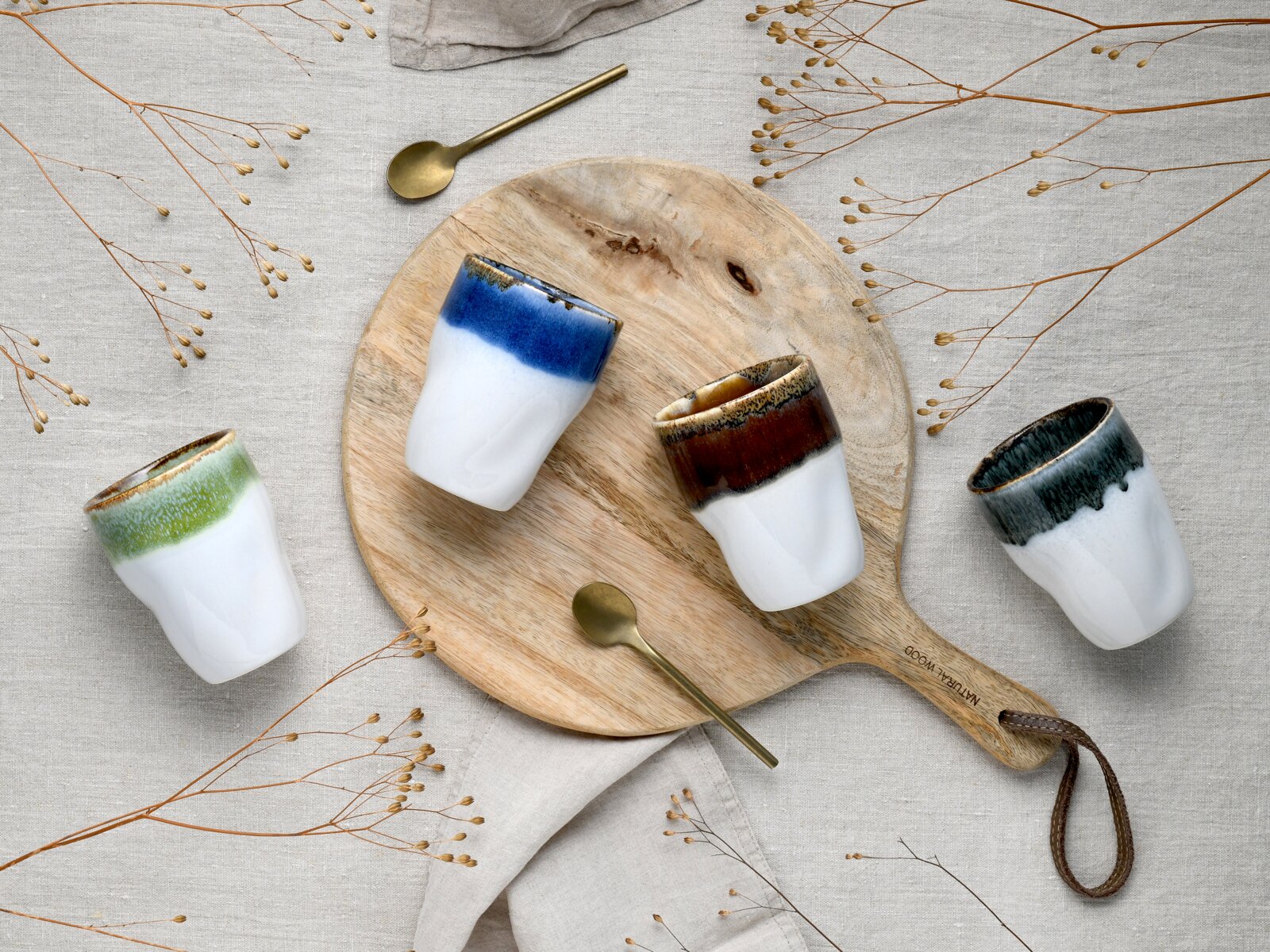 Drei kleine Keramikbecher in Weiß mit farbigem Rand (Blau, Braun und Dunkelgrün) auf rundem Holzbrett mit goldenen Löffeln und Trockenblumen auf Leinentuch, Draufsicht von oben