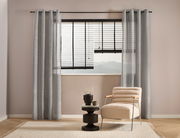 Grauer Ösenschal 135x245cm vor einem Fenster mit Blick auf Berge, im Vordergrund ein beiger Sessel und ein kleiner Tisch, Perspektive von vorne.