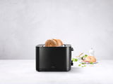 Schwarzer Toaster mit zwei Scheiben Brot, seitliche Perspektive, auf einem hellen Hintergrund mit Bagel und Gewürzen im Hintergrund.