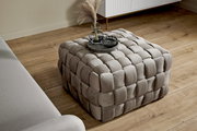 Quadratischer, gepolsterter Hocker mit geflochtenem Design in beige, von oben betrachtet, auf einem Holzboden neben einem Sofa.