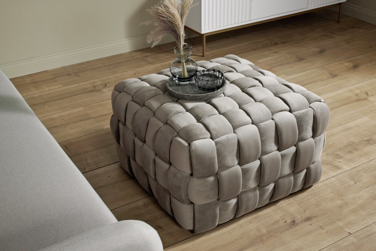Vierkante, gestoffeerde poef met gevlochten patroon in beige, gezien van bovenaf, op een houten vloer naast een bank.