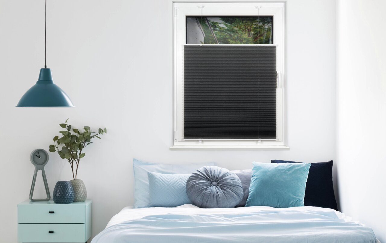 Schwarzes Plissee am Fenster in einem modern eingerichteten Schlafzimmer mit blauen Kissen und einer Hängelampe. Perspektive von vorne.