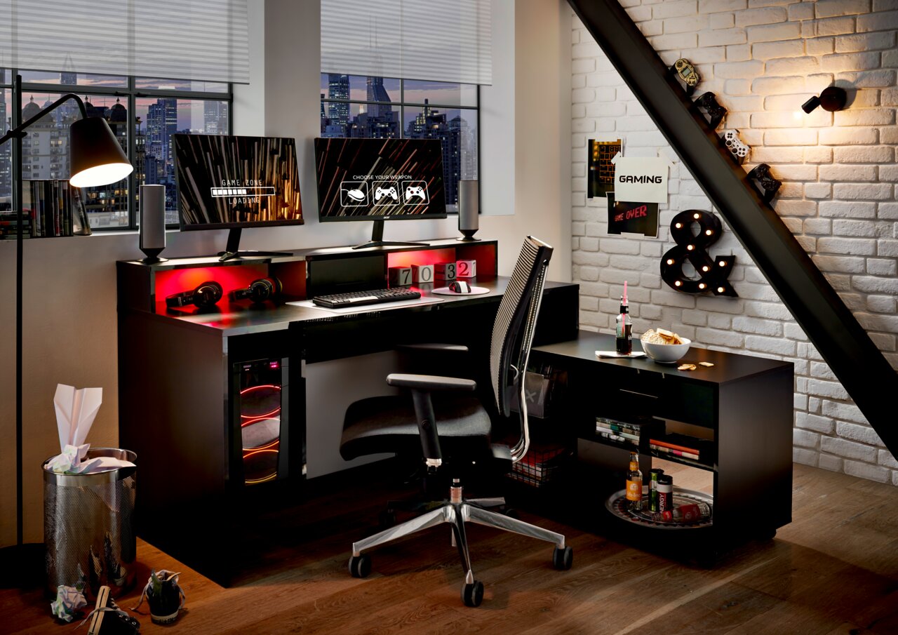 Moderner Gaming-Schreibtisch mit zwei Monitoren, beleuchtetem PC-Gehäuse und ergonomischem Stuhl in einem stilvollen Raum mit Stadtblick, aus seitlicher Perspektive.