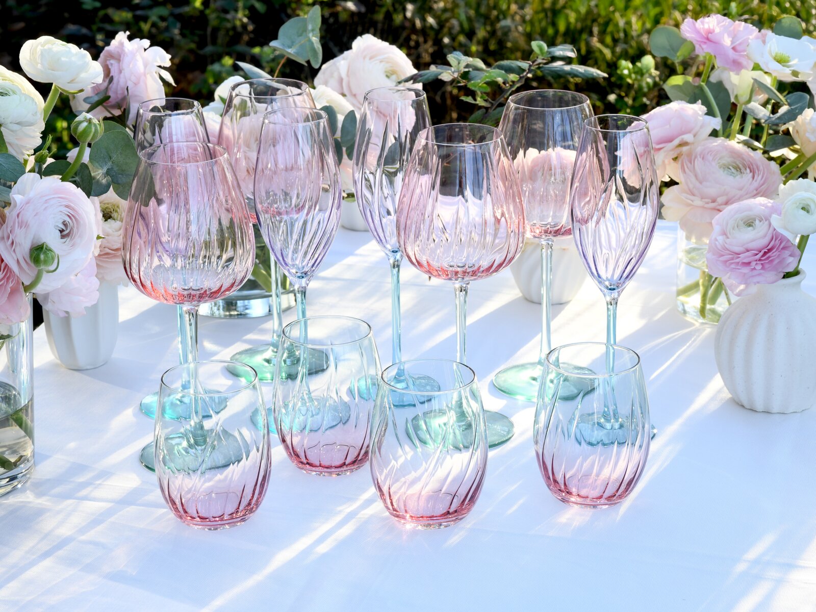 Schräg von vorne fotografiertes Set aus facettierten Glas-Trinkgläsern und Weingläsern mit zartem Farbverlauf in Rosa und Mint auf weißer Tischdecke, dekoriert mit pastellfarbenen Blumen im Garten.