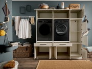 Frontalansicht eines modernen Hauswirtschaftsraums mit offenem, weißem Einbauschrank für Waschmaschine und Trockner in Schwarz, mit Regalfächern, Schubladen und geöffneter Seitentür, daneben Kleiderstange und Wäschekörbe.