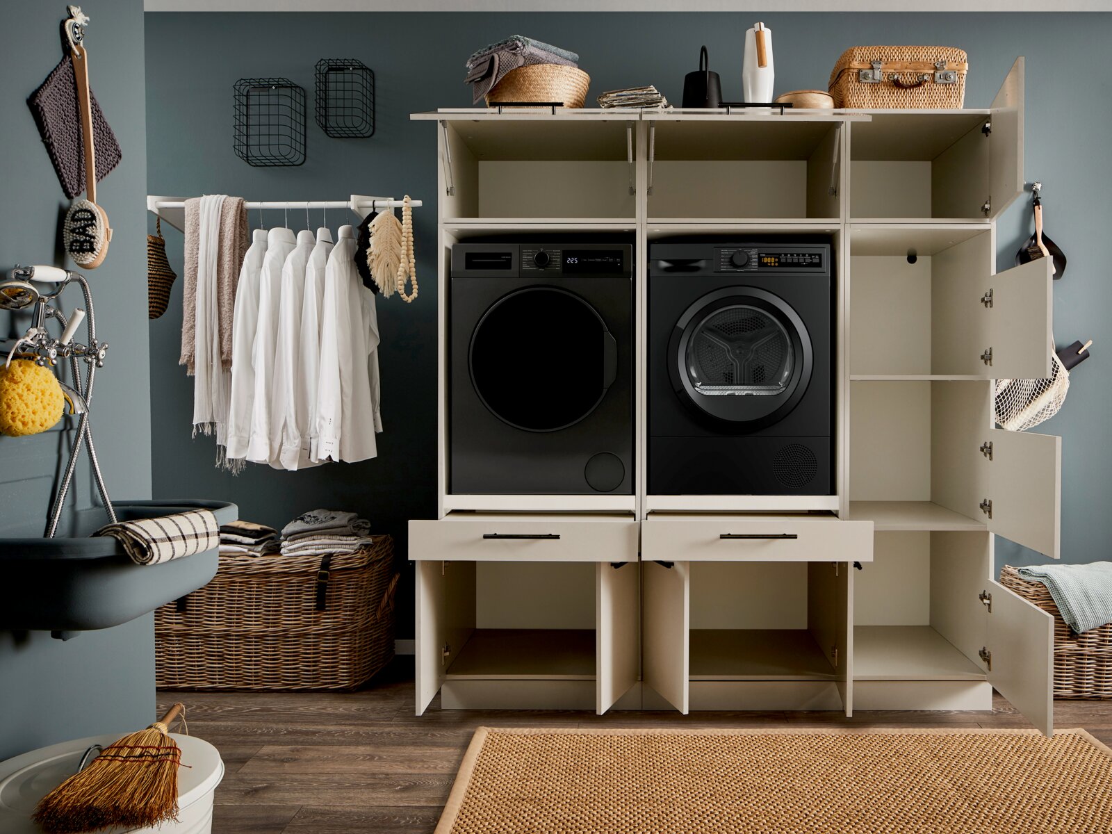 Frontalansicht eines modernen Hauswirtschaftsraums mit offenem, weißem Einbauschrank für Waschmaschine und Trockner in Schwarz, mit Regalfächern, Schubladen und geöffneter Seitentür, daneben Kleiderstange und Wäschekörbe.