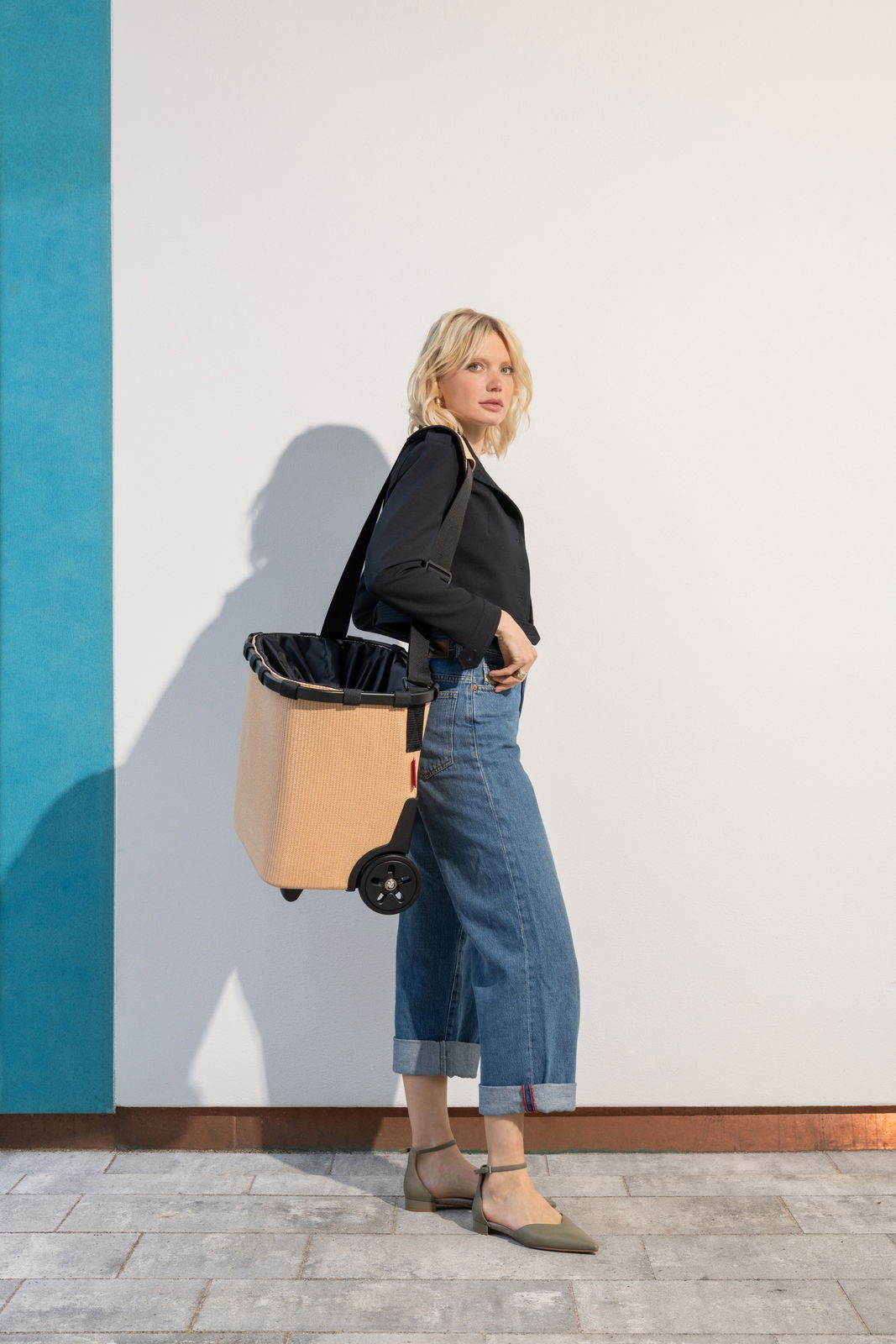 Reisenthel Carrycruiser Raffia Black Frau in seitlicher Perspektive mit einem beigen carrycruiser auf Rollen, der über der Schulter getragen wird.