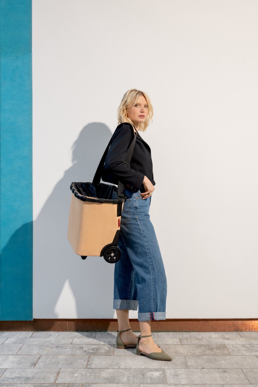 Vrouw in zijaanzicht met een beige carrycruiser op wieltjes, die over de schouder wordt gedragen.