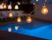 Lichterkette mit Glühbirnen über einem beleuchteten Pool bei Nacht, seitliche Perspektive