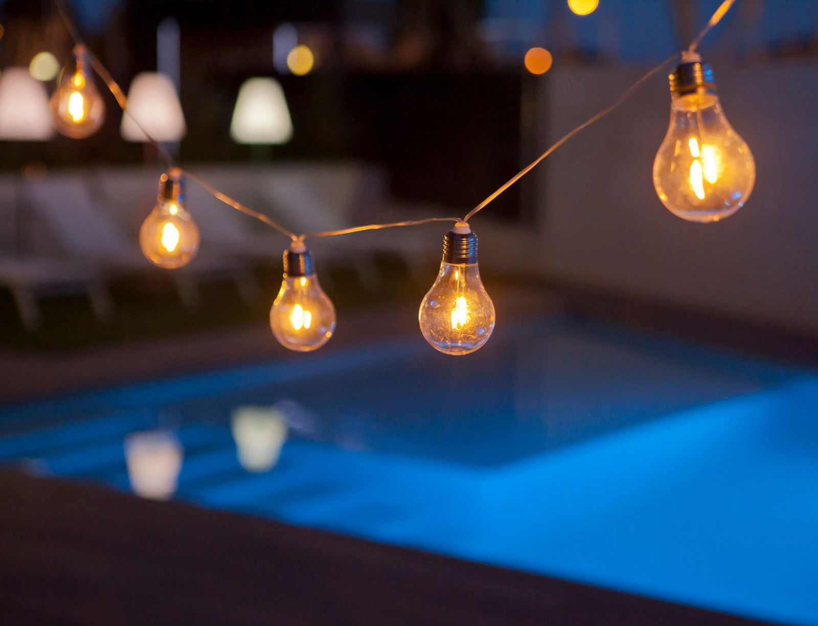 Lichterkette mit Glühbirnen über einem beleuchteten Pool bei Nacht, seitliche Perspektive