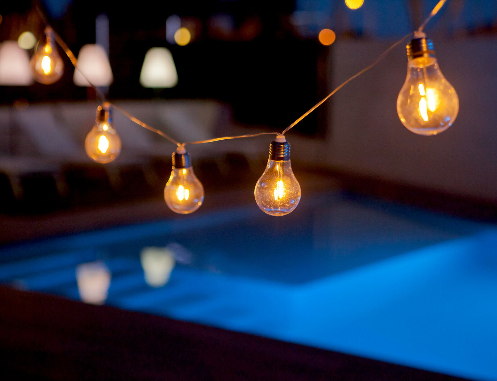 Lichterkette mit Glühbirnen über einem beleuchteten Pool bei Nacht, seitliche Perspektive