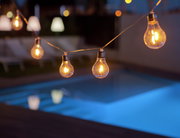 Lichterkette mit Glühbirnen über einem beleuchteten Pool bei Nacht, seitliche Perspektive