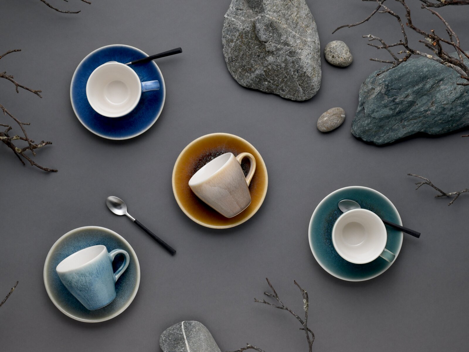 Draufsicht (Flatlay) auf vier farbige Kaffeetassen mit Untertassen und Löffeln auf grauem Hintergrund, dekoriert mit Steinen und Zweigen.