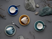 Draufsicht (Flatlay) auf vier farbige Kaffeetassen mit Untertassen und Löffeln auf grauem Hintergrund, dekoriert mit Steinen und Zweigen.